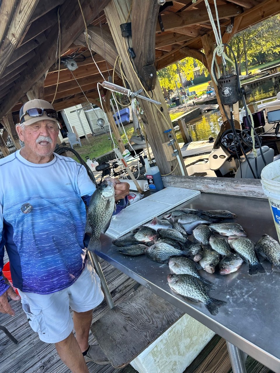 A fisherman holding a crappie beside a dockside cleaning table covered with the catch