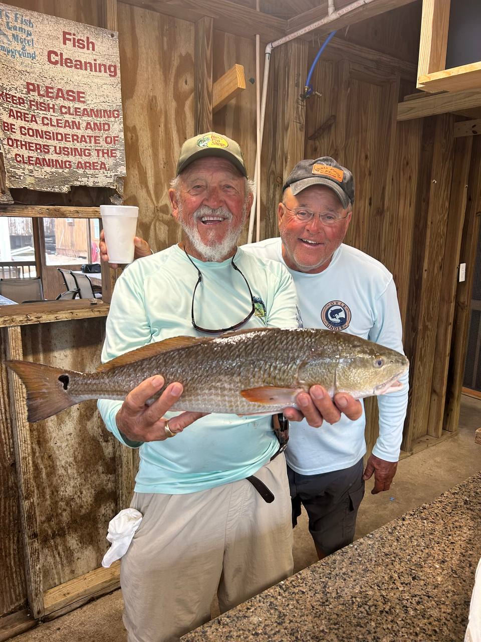 A smiling fisherman holding a large redfish in the cleaning room with Captain Jim beside him
